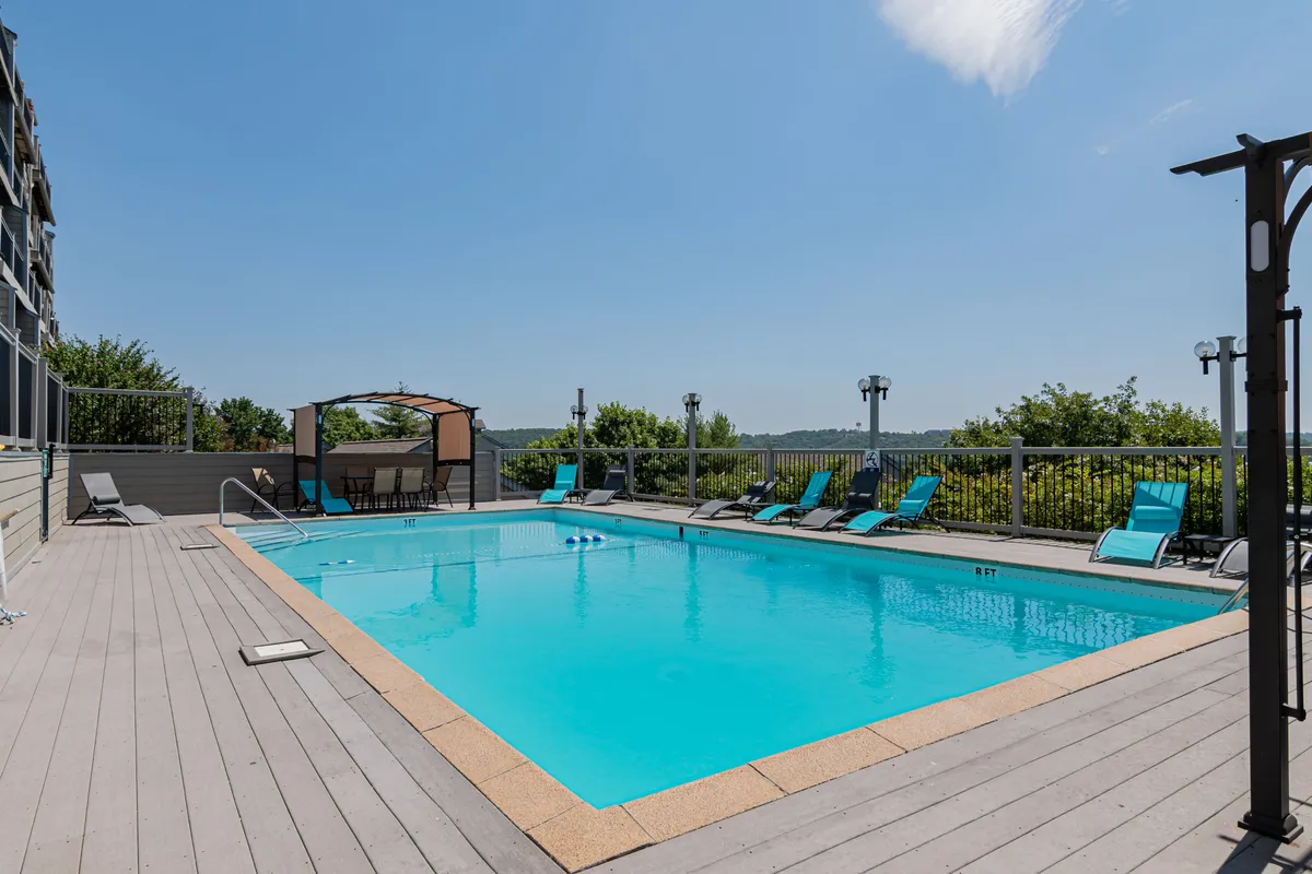 A sparkling outdoor swimming pool is surrounded by comfortable lounge chairs and a shaded canopy seating area, set against a backdrop of trees and blue skies. The scene suggests a relaxing and fun environment where families can unwind, swim, and socialize after a day at nearby attractions.