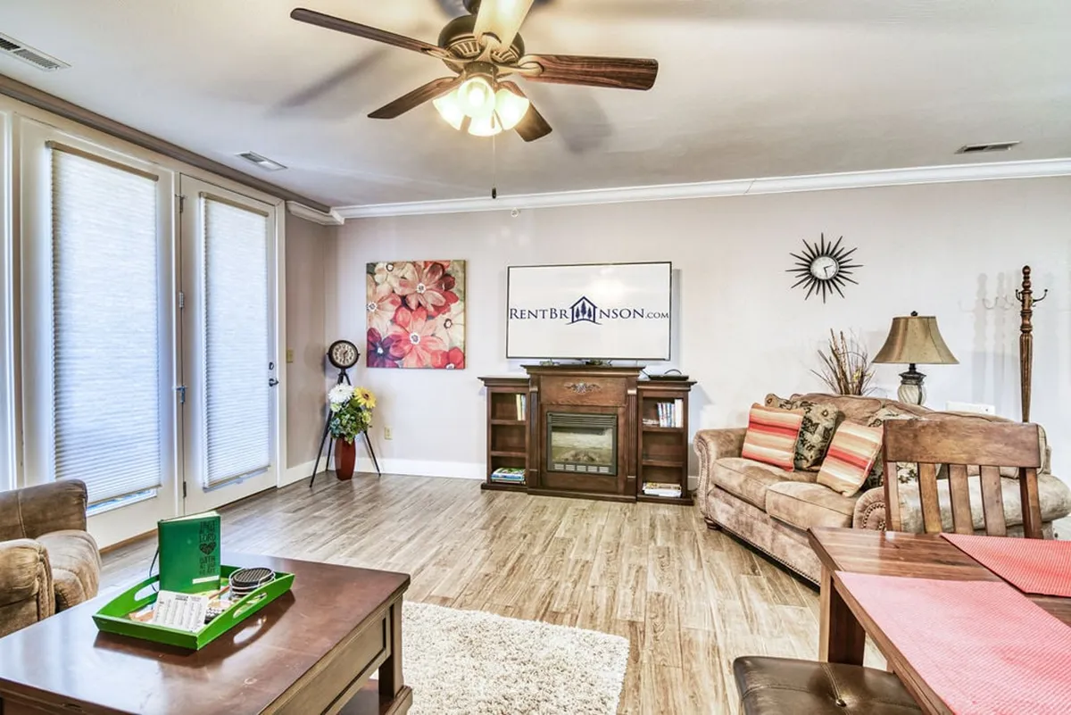 The image features a stylish, inviting living area with a plush sofa, modern coffee table, vibrant artwork, a large television, and expansive windows that fill the space with natural light. A cozy electric fireplace anchors the room, creating a warm and welcoming atmosphere perfect for relaxing or socializing.