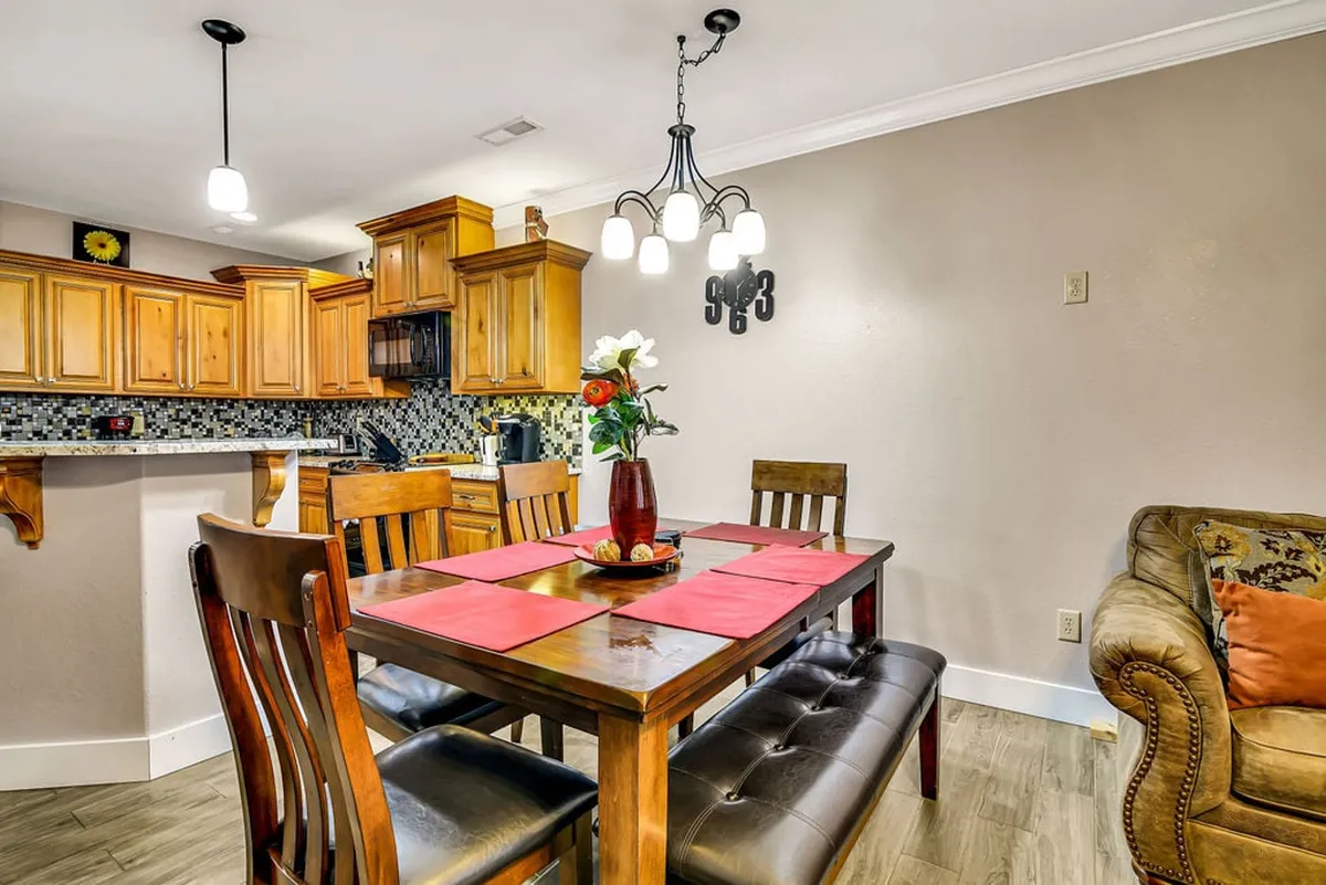 A softly lit dining area connected to the kitchen, with a prominent dining table set beneath a chandelier and decorative floral accents. The scene hints at shared meals, celebrations, and leisurely breakfasts with views into the heart of the home.