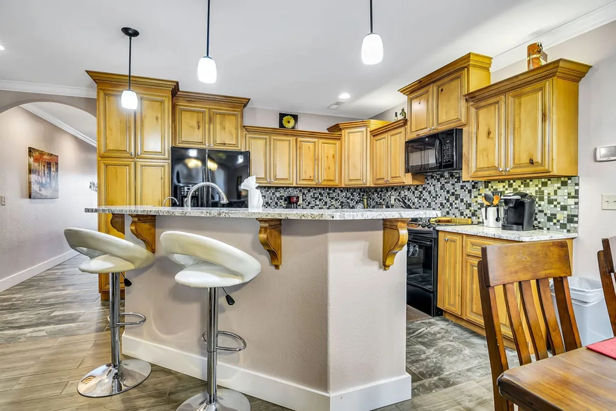 A bright, stylish kitchen featuring a spacious island with granite countertop, wooden cabinetry, and bar stools. The open layout encourages conversation and easy movement, while modern appliances are visible in the background, reflecting a welcoming, functional cooking space perfect for gatherings.